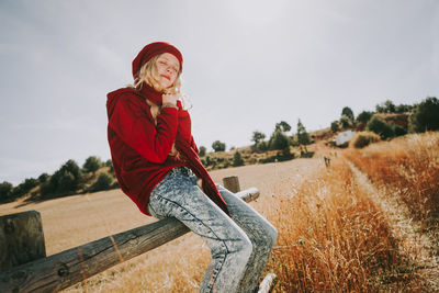 Woman sitting on railing in field against sky