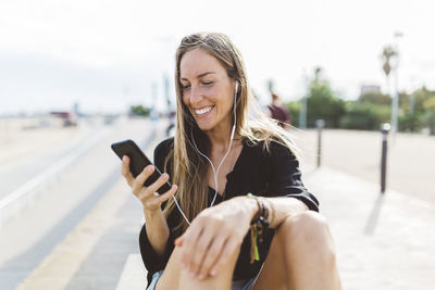 Smiling young woman with cell phone and earbuds on waterfront promenade