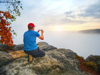 Hiker takes selfie photo, fall nature adventure. man sit on stone on mountain sumit daybreak horizon