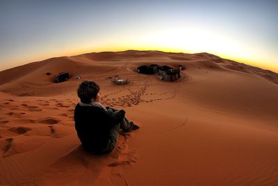 Rear view of woman sitting on sand at desert against clear sky