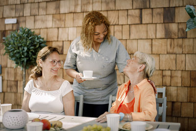 Happy young female nurse talking with senior women sitting at dining table