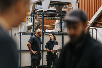 Mature man with arms akimbo standing next to female colleague while discussing strategy in factory