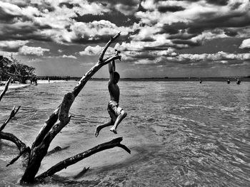Shirtless boy hanging on driftwood over sea against sky