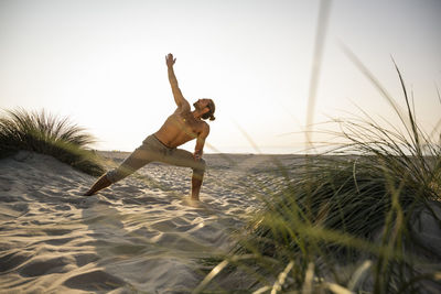 Shirtless young man practicing triangle position yoga at beach against clear sky during sunset