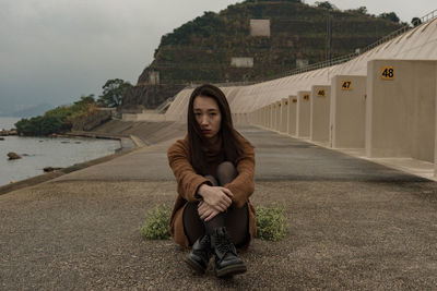 Portrait of young woman sitting on footpath by sea