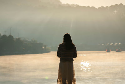 Rear view of woman standing by lake against sky during sunset
