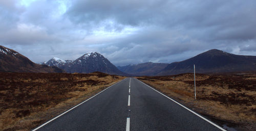 Empty road by mountains against cloudy sky