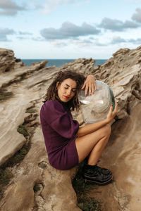 Side view of female sitting on rocky ground and hugging glass cosmonaut helmet while pretending being spaceman