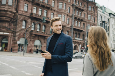 Smiling businessman with phone looking at female coworker while standing in city