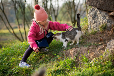 Full length of girl petting cat while crouching on grassy field