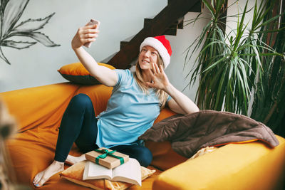 A smiling girl in a christmas hat sits on the sofa and communicates via video link with friends. 