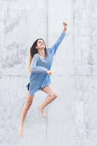 Happy young woman with arm raised jumping against concrete wall