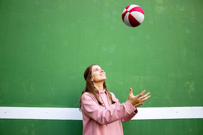 Smiling young woman playing with ball against wall