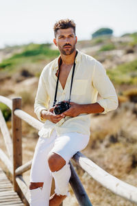 Young man holding camera sitting on railing against sky