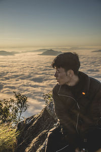 Young man looking away while sitting on mountain against clear sky during sunset