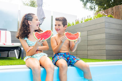 Two funny kids eating watermelon at the the edge of the pool on a summer day