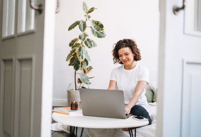 View from doors on young brunette woman in white t-shirt using laptop sitting on bed at home