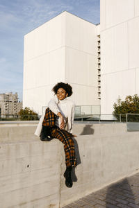 Carefree young woman with afro hair sitting on retaining wall against buildings in city