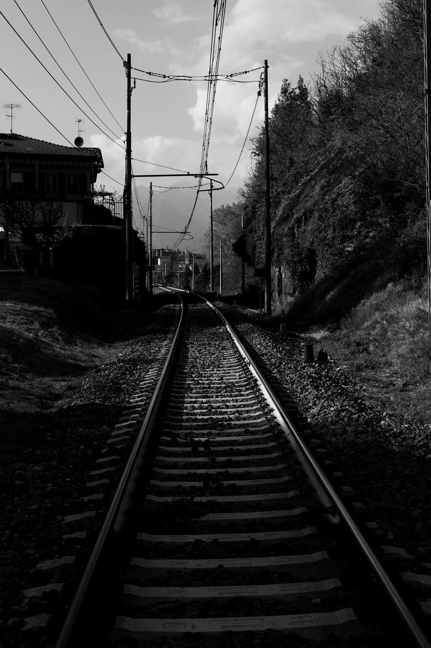 RAILROAD TRACKS AMIDST PLANTS AND ELECTRICITY PYLON AGAINST SKY