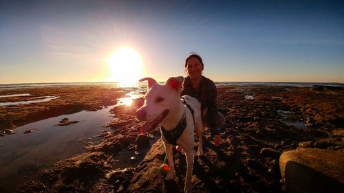 Smiling woman with dog at beach against sky during sunset