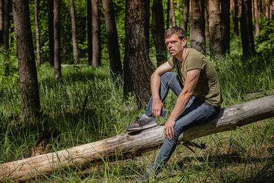 A man in a forest sits leaning against a tree, with headphones in her ears listening to music. 