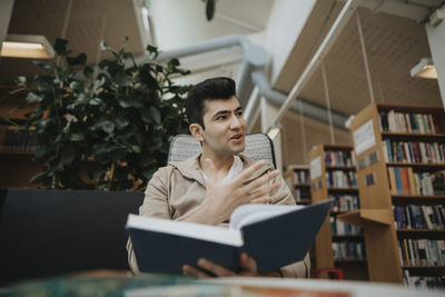 Male student gesturing while holding book in library