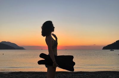 Side view of woman standing at beach against sky during sunset