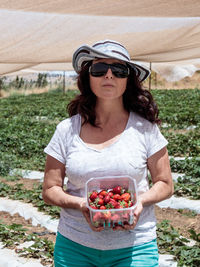 Portrait of woman holding strawberries in container while standing at farm during sunny day