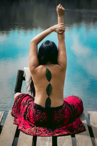 Rear view of shirtless woman with leaves on back sitting at pier by sea