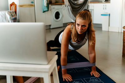 Sportswoman performing exercise with resistance bands while watching tutorials on laptop and working out at home