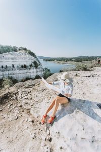 Woman reading map while sitting on rock formation against clear sky