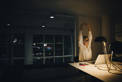 Exhausted businesswoman stretching with arms raised while working late in illuminated coworking office