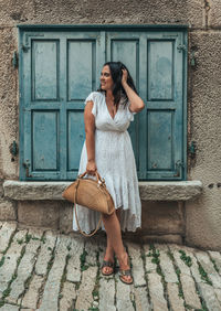 Full length portrait of beautiful young woman standing in front of blue window shutters in old town