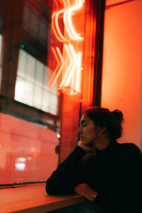 Side view of gorgeous young ethnic woman looking out window with red lightening on the background leaning on hand and dreaming