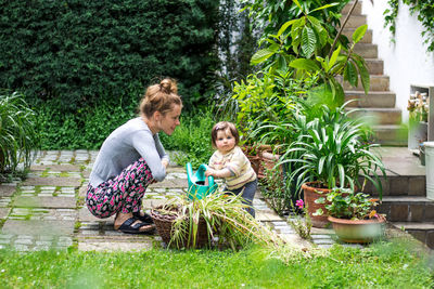 Young mother and her toddler child are doing garden work with a watering can in the yard