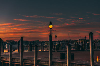 Illuminated street light by buildings against sky during sunset