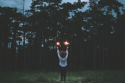 Rear view of woman holding illuminated sparklers in forest during dusk