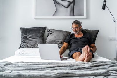 Smiling middle aged male entrepreneur sitting on bed and typing on laptop while working remotely on online project