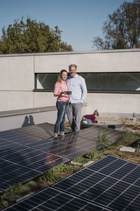 Smiling woman holding tablet pc standing with man amidst solar panel on sunny day