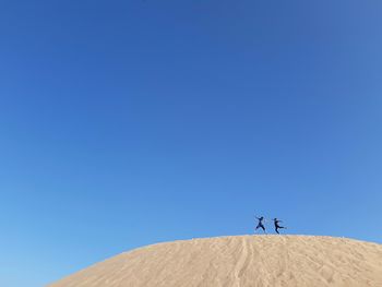 Low angle view of boys jumping on sand dune at desert against clear blue sky