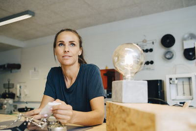 Thoughtful female technician looking away while sitting at workbench in creative office