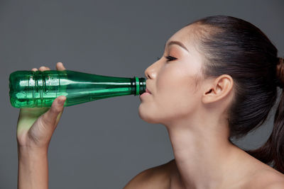 Close-up of young woman drinking water from bottle against gray background