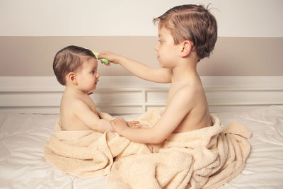 Side view of shirtless boy combing brother hair on bed at home