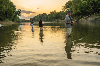 Friends enjoying fishing in lake against sky during sunset