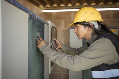 Side view of female engineer programming fuse box through smart phone at power station