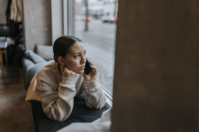 Businesswoman talking over mobile phone while looking out through window lying on seat at office