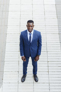 From above confident adult african american male entrepreneur in elegant blue suit with tie looking at camera while standing against blurred urban background of modern city