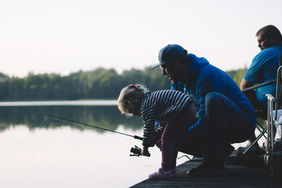 Side view of daughter and father fishing in lake against sky