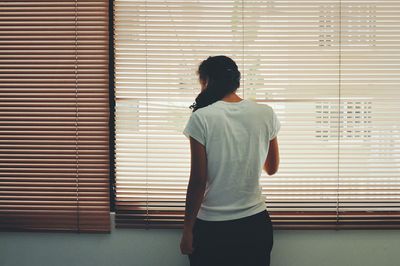 Rear view of woman standing by window blinds at home