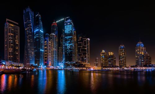 Illuminated modern buildings by river against sky at night